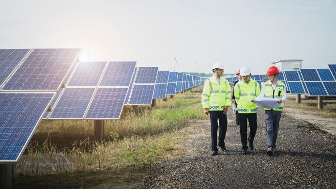 Dekarbonisierung: Person mit Sicherheitsweste und Schutzhelm, steht vor einer Reihe von Windrädern und schaut nach rechts. Die Sonne geht im Hintergrund unter.