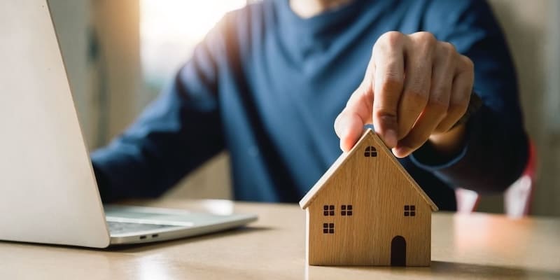 Person arbeitet am Laptop und hält ein kleines Holzhaus-Modell als Symbol für Immobilienkauf und Beratung – Dr. Klein Baufinanzierung AB