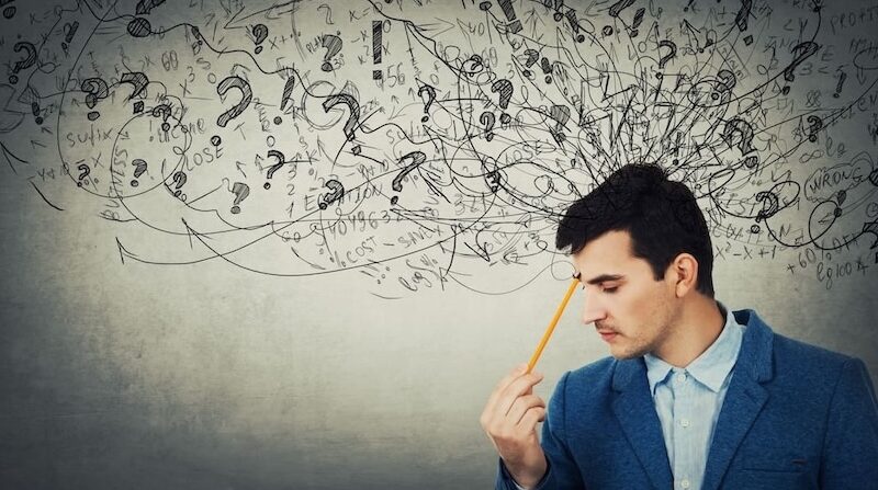 Symbolbild für einen überladenen Geist: Ein nachdenklicher Mann hält einen Stift an die Stirn, während wirre Gedanken, Skizzen und Fragezeichen seinen inneren Stress und kognitive Überforderung verdeutlichen.