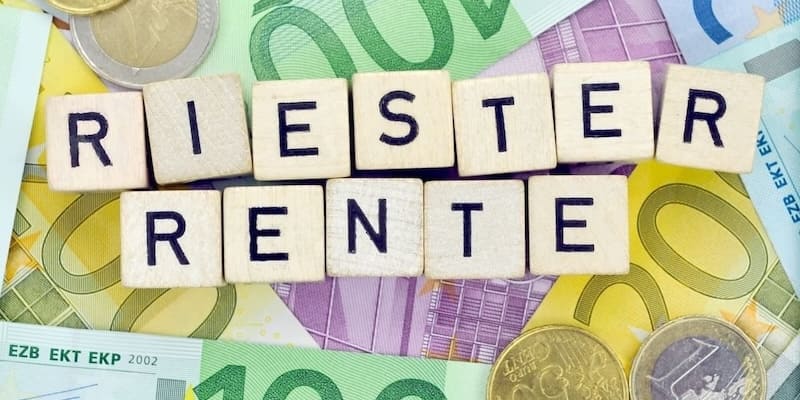 Schriftzug „Riester Rente“ aus Holzklötzen auf farbigen Euro-Scheinen und Münzen. Neutrales Symbolbild für Riester Rente für Berufseinsteiger.